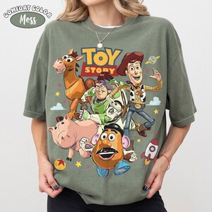 May include: A Comfort Colors Moss green t-shirt featuring the Toy Story logo and characters. The graphic includes Woody, Buzz Lightyear, Mr. Potato Head, Hamm, and Bullseye. The shirt has a crew neck and short sleeves.