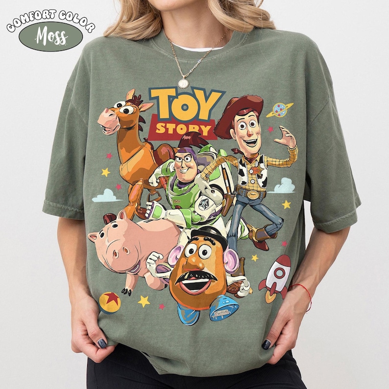 May include: A Comfort Colors Moss green t-shirt featuring the Toy Story logo and characters. The graphic includes Woody, Buzz Lightyear, Mr. Potato Head, Hamm, and Bullseye. The shirt has a crew neck and short sleeves.
