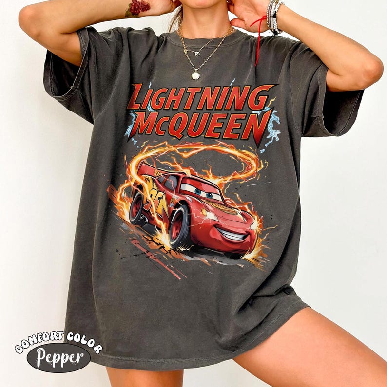 Op de afbeelding: Donkergrijs t-shirt met Lightning McQueen uit de film Cars. De afbeelding toont de rode raceauto met vlammen en de tekst "Lightning McQueen" in rood en geel. Het shirt is gemarkeerd met "Comfort Color Pepper".