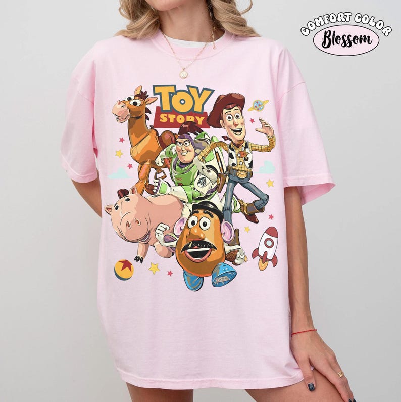 May include: A light pink Comfort Colors t-shirt featuring a graphic of Toy Story characters, including Woody, Buzz Lightyear, and Mr. Potato Head. The shirt has the words "Toy Story" printed in a colorful font. A rocket ship and stars are also included in the design.