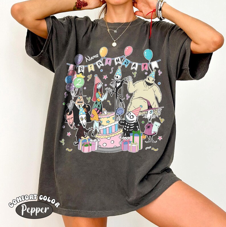 Op de afbeelding: Donkergrijs Comfort Colors T-shirt met een kleurrijke afbeelding van personages uit The Nightmare Before Christmas die een verjaardag vieren. Het ontwerp bevat een taart, ballonnen en het woord "BIRTHDAY".
