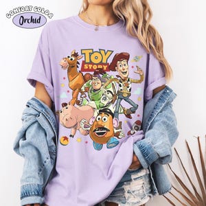 May include: A light purple Comfort Colors t-shirt featuring a graphic of characters from the animated film "Toy Story." The design includes Woody, Buzz Lightyear, and other characters. The shirt has the text "TOY STORY" in yellow and red.