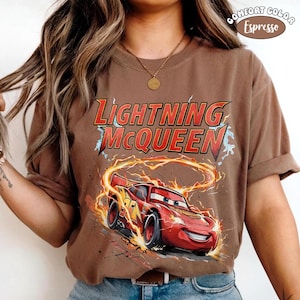 Op de afbeelding: Bruin T-shirt met Lightning McQueen uit de film Cars. Het ontwerp bevat de tekst "Lightning McQueen" in rood en geel, en een afbeelding van de rode raceauto omringd door vlammen en bliksem.