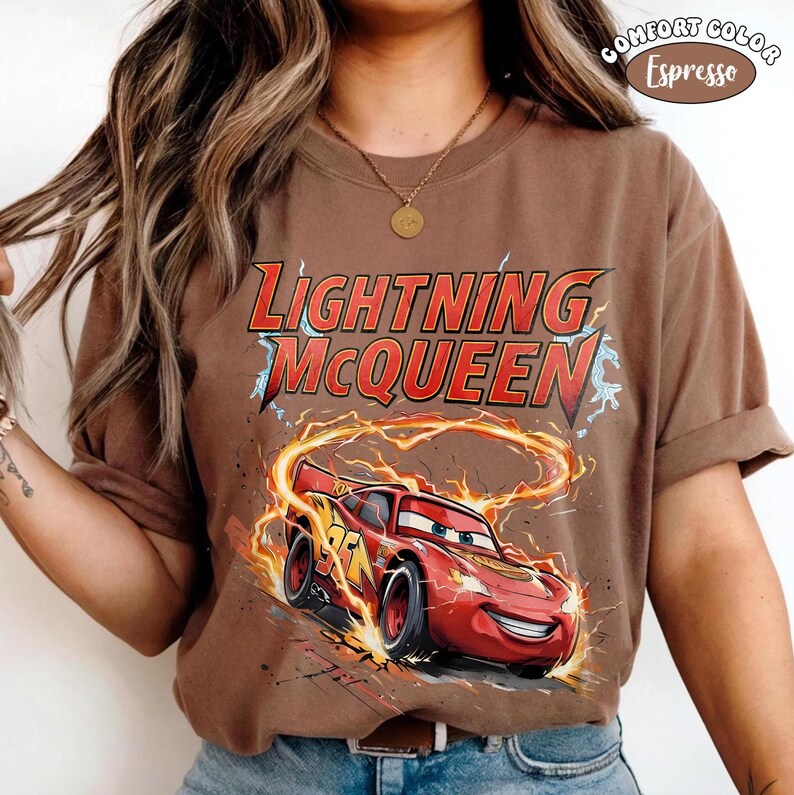 Op de afbeelding: Bruin T-shirt met Lightning McQueen uit de film Cars. Het ontwerp bevat de tekst "Lightning McQueen" in rood en geel, en een afbeelding van de rode raceauto omringd door vlammen en bliksem.