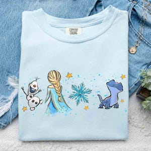 K&ouml;nnte beinhalten: Ein hellblaues T-Shirt mit Rundhalsausschnitt und einem Grafikdruck, der einen Schneemann, eine blonde Figur mit Zopf, eine blaue Schneeflocke und eine kleine blaue Eidechse zeigt, akzentuiert durch gelbe Sterne und blaue Farbspritzer.