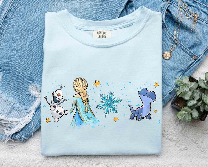 K&ouml;nnte beinhalten: Ein hellblaues T-Shirt mit Rundhalsausschnitt und einem Grafikdruck, der einen Schneemann, eine blonde Figur mit Zopf, eine blaue Schneeflocke und eine kleine blaue Eidechse zeigt, akzentuiert durch gelbe Sterne und blaue Farbspritzer.