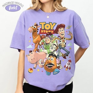 May include: A Comfort Colors violet t-shirt featuring a colorful graphic of Toy Story characters. The design includes Woody, Buzz Lightyear, and other characters from the animated film. The shirt has a crew neck and short sleeves.