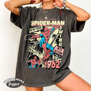 Puede incluir: Camiseta gris oscuro con un gr&aacute;fico de estilo c&oacute;mic vintage de Spider-Man balance&aacute;ndose por la ciudad de Nueva York. El dise&ntilde;o incluye el texto "THE AMAZING SPIDER-MAN NEW YORK 1962" y "SWING INTO ACTION". La camiseta tiene una etiqueta Comfort Color Pepper.
