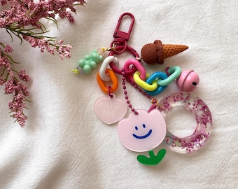 May include: A whimsical keychain with a burgundy clasp and a variety of charms. Charms include a pink flower with a smiley face, a heart with "HUG ME" text, a brown ice cream cone, and a clear ring with pink glitter.