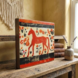May include: A wooden block featuring a red horse illustration with orange floral accents. The artwork has a cream background with black and red borders. The piece is displayed on a rustic wooden table.