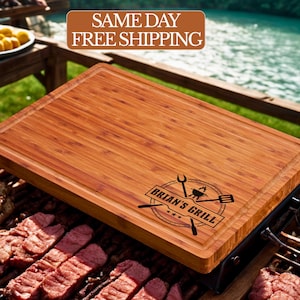 May include: A rectangular bamboo cutting board with a juice groove and the text "Brian's Grill" is displayed over a grill with raw meat. The board is brown with a dark engraved logo. The background shows a lake and wooden table.
