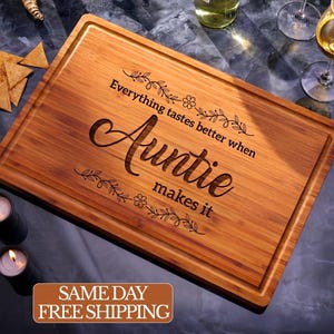 May include: A rectangular wooden cutting board with a juice groove and the engraved text "Everything tastes better when Auntie makes it." The board is surrounded by food and drinks, including wine glasses and tortilla chips.