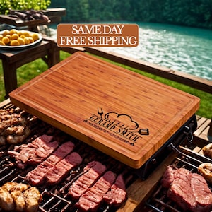 May include: A rectangular bamboo cutting board with a juice groove and the text "GERARD SMITH BEST COOK" in black. The board is placed on a grill with grilling food. The background shows a lake and wooden furniture.