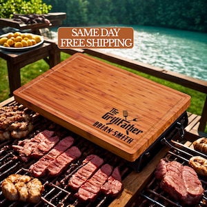 May include: A rectangular bamboo cutting board with a juice groove. The board has the text "The GrillFather" and "BRIAN SMITH" engraved on it. The board is on a grill with cooked meat.