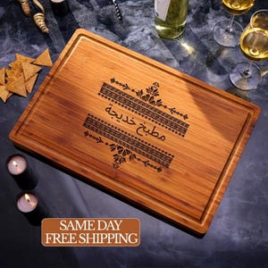 May include: A rectangular bamboo cutting board featuring a juice groove along the perimeter and a dark laser-engraved design in the center. The engraving includes intricate geometric patterns and the Arabic text "مطبخ خديجة".