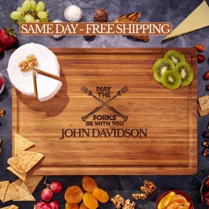 May include: A rectangular bamboo cutting board with a juice groove, engraved with two crossed forks and the text MAY THE FORKS BE WITH YOU and JOHN DAVIDSON. The board is surrounded by various food items including grapes, cheese, and crackers.
