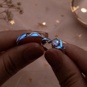 Könnte beinhalten: Zwei silberfarbene Ringe mit blauem Leuchteffekt. Ein Ring hat eine glatte Schiene mit leuchtenden Akzenten, während der andere einen zentralen runden Stein, flankiert von zwei kleinen klaren Steinen und einem leuchtend blauen Walflossenmotiv, zeigt.