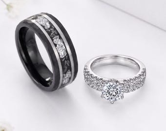 Puede incluir: Un anillo negro con una incrustación central texturizada con fragmentos de plata metálica y un anillo plateado con una piedra central de corte redondo y una banda incrustada con pequeñas piedras claras a ambos lados, presentados juntos.