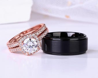 Puede incluir: Un conjunto de dos anillos de boda. El anillo de compromiso es de oro rosa con una piedra grande, redonda y transparente rodeada de piedras más pequeñas. La banda de boda es negra y tiene un acabado cepillado.