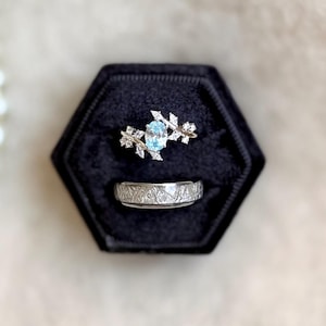 May include: A silver-toned ring featuring an oval light blue center stone surrounded by small clear stones in a leaf-like arrangement, positioned above a second silver-toned band with a textured, cross-hatched metallic surface pattern.
