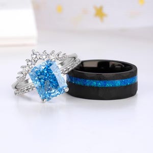 May include: Two rings: a silver engagement ring with a large, rectangular, light blue gemstone and a black band ring with a blue opal inlay. The engagement ring has a halo of small diamonds around the main stone.