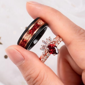 Op de afbeelding: Een paar trouwringen. De eerste ring is een zwarte en roségouden band met rode en gouden glitter ingebed in het roségoud. De tweede ring is een roségouden band met een peervormige rode edelsteen omgeven door een halo van kleinere witte edelstenen.