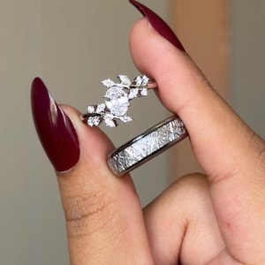 Könnte beinhalten: Ein silberfarbenes Ring-Set bestehend aus einem Ring mit einem oval geschliffenen Mittelstein, der von blattförmigen Akzenten umgeben ist, und einem zweiten Ring mit einer texturierten, kreuzschraffierten Oberfläche.