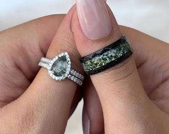 Op de afbeelding: Een peervormige verlovingsring met een groene mosagaat-middelsteen omringd door een halo van kleine diamanten. De ring wordt aan de linkerhand gedragen, naast een zwart en groene band met een mosagaat-inleg.