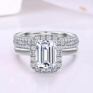 Puede incluir: Un anillo de compromiso de plata con una gran piedra central de corte esmeralda, transparente, rodeada por un halo de diamantes más pequeños. La banda también está engastada con diamantes más pequeños.