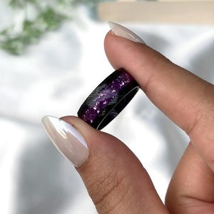 May include: A black ring with purple glitter embedded in the resin. The ring is shown on a finger.