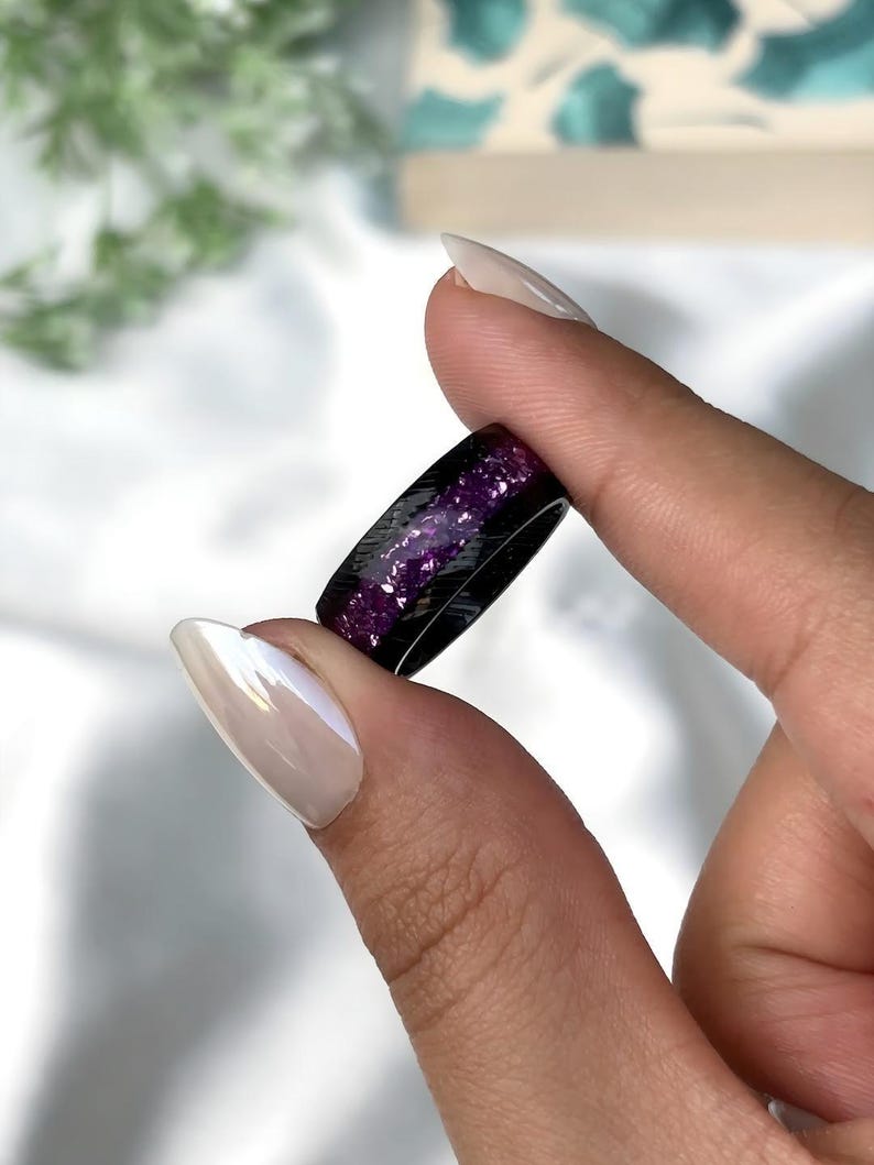 May include: A black ring with purple glitter embedded in the resin. The ring is shown on a finger.