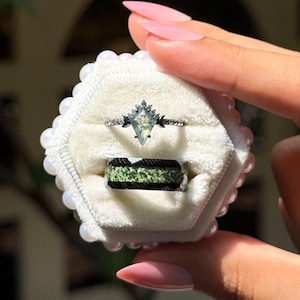 Puede incluir: Dos anillos presentados en una caja blanca con borde de perlas. El anillo superior tiene una piedra verde claro en forma de diamante con una banda plateada. El anillo inferior tiene una banda negra con una incrustación verde texturizada.