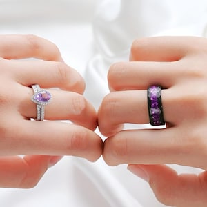 May include: Two rings are displayed, one on each hand. The ring on the left is silver with a pear-shaped purple gemstone surrounded by smaller stones. The ring on the right is black with purple and white accents. The background is a soft white fabric.
