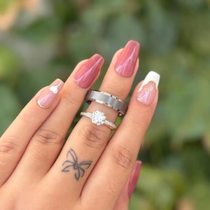 Peut inclure: Deux bagues sont présentées sur un doigt. La bague du haut est argentée avec une bande texturée et une bande intérieure dorée. La bague du bas est dorée avec une grosse pierre ronde et transparente et de petites pierres autour de la bande. Les ongles sont peints avec du vernis à ongles rose et blanc.