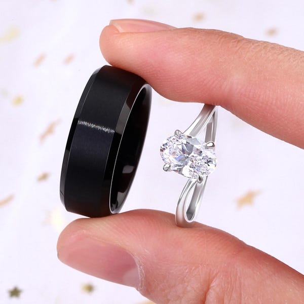 May include: A pair of wedding bands. The woman's ring features a large oval-shaped diamond set in a silver band. The man's ring is a black band with a bevelled edge.
