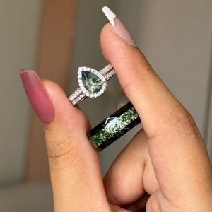 May include: Two rings are displayed. One is a silver band with a pear-shaped green stone surrounded by a halo of small clear stones. The other is a black band with a green and white speckled design.