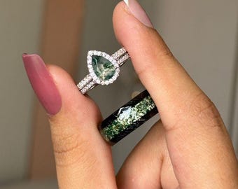May include: Two rings are displayed. One is a silver band with a pear-shaped green stone surrounded by a halo of small clear stones. The other is a black band with a green and white speckled design.