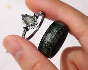 Puede incluir: Se muestran dos anillos. Uno es un anillo negro con una piedra de talla cuadrada, de color verde y pequeñas piedras blancas. El otro es una banda negra con una incrustación estampada en verde y blanco. Los anillos están sostenidos por los dedos de una persona.