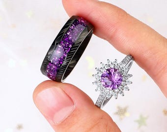 Op de afbeelding: Een paar trouwringen met een zwart en paars ontwerp. De ring aan de linkerkant heeft een zwarte band met een rij paarse stenen. De ring aan de rechterkant heeft een zilveren band met een grote paarse steen omringd door kleinere witte stenen.