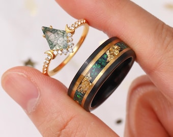 May include: Two rings are displayed. One is gold with a green, kite-shaped stone and small diamonds. The other is a black band with gold flakes and a green inlay. The rings are held up by a person's fingers.