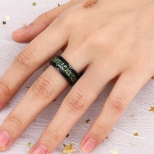 May include: A black and green ring with a textured design. The ring is worn on a person's finger.