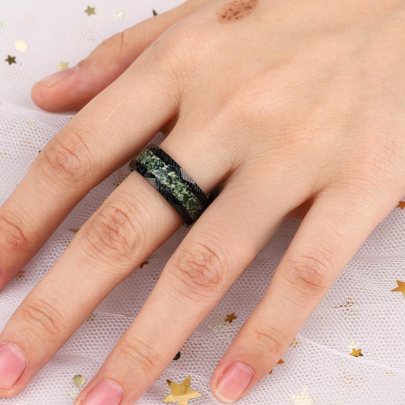 May include: A black and green ring with a textured design. The ring is worn on a person's finger.