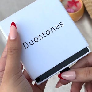 May include: A white box with the text "Duostones" printed in black. The box is being held by a hand with red and white nail polish.
