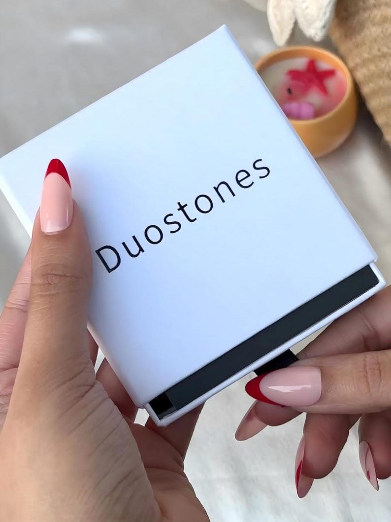 May include: A white box with the text "Duostones" printed in black. The box is being held by a hand with red and white nail polish.