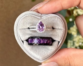 May include: A heart-shaped gray velvet ring box holds three rings. The top ring is a pear-shaped purple gemstone with a halo of smaller stones. The second ring is a band with small stones. The third ring is a black band with purple flecks.