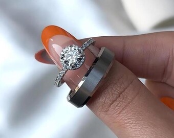 Puede incluir: Dos anillos plateados, uno con una piedra grande, redonda y transparente rodeada de piedras más pequeñas, y una banda más ancha con un acabado mate. Los anillos se muestran en un dedo con esmalte de uñas naranja.