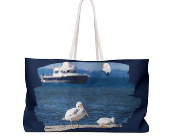 May include: Navy blue tote bag featuring a scenic image of white pelicans near a body of water with a boat in the background. The bag has white rope handles and is designed for carrying items.