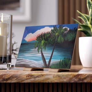 Könnte beinhalten: Ein quadratisches Gemälde einer tropischen Strandszene mit zwei Palmen, blauem Wasser und einer rosa-weißen Wolke. Das Gemälde wird auf einer Holzstaffelei ausgestellt. Ein lebendiges Kunstwerk für die Inneneinrichtung.