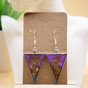 Peut inclure: Boucles d'oreilles triangulaires avec des montures argentées. Elles présentent un motif marbré violet et marron, avec des touches de turquoise. Elles sont présentées sur une carte marron avec le texte "Ecnelis".