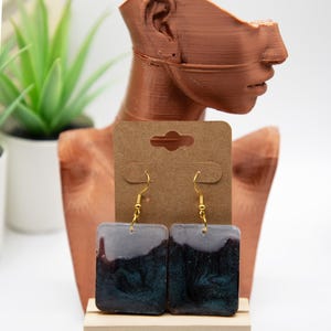 May include: Square resin earrings with gold-coloured hooks. The earrings have a dark teal and grey marbled design. The earrings are displayed on a brown earring card and a wooden stand. The word "Ecnelis" is in the upper right corner.